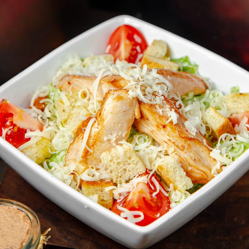 Chicken Caesar salad with grilled chicken, croutons, tomatoes, and shredded cheese in a white bowl.
